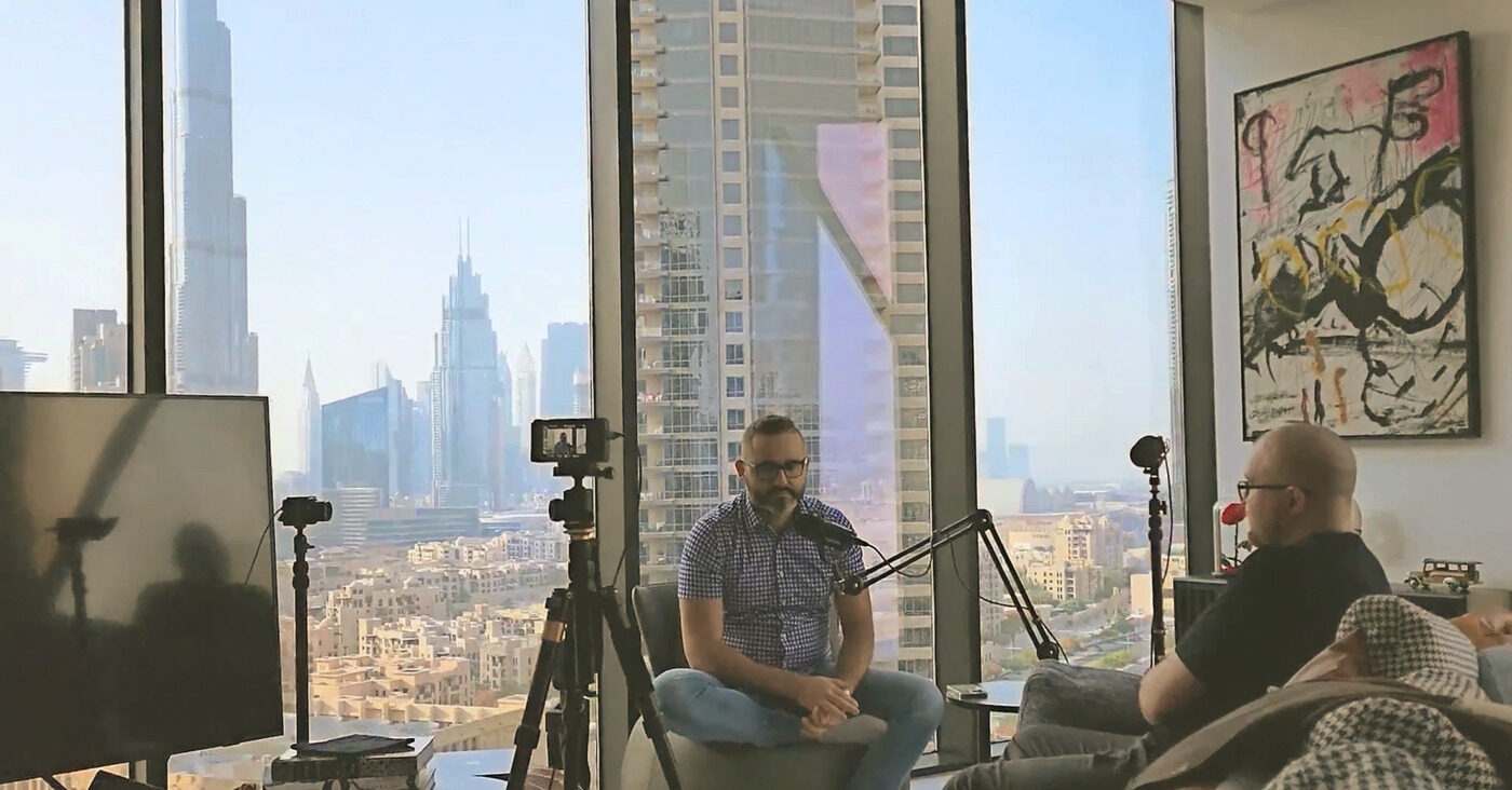 Tom recording the BuddyBoss podcast with Graham in his living room, floor-to-ceiling windows and the Dubai skyline with the Burj Khalifa behind them.